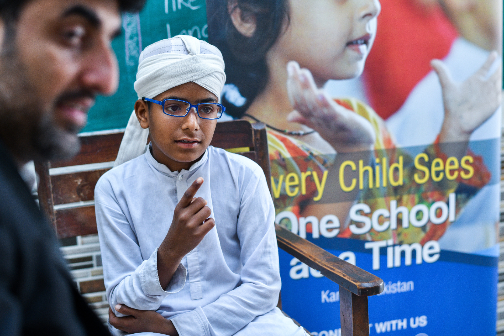 Pakistani boy in new blue glasses speaks with a teacher in front of an OSaaT 'Every Child Sees, One School at a Time' Karachi banner