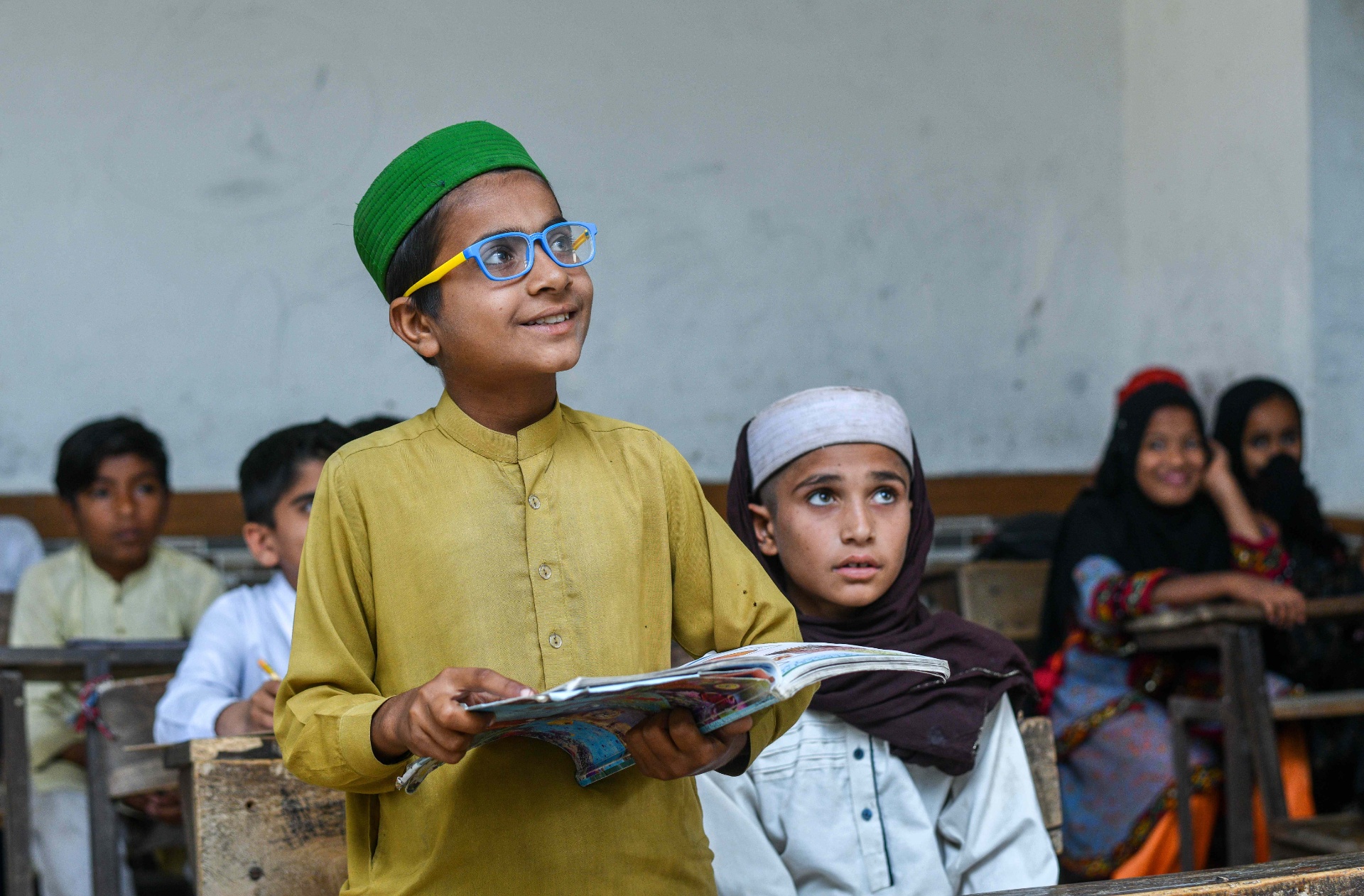 Pakistani student in blue glasses stands and reads aloud from his book in a classroom