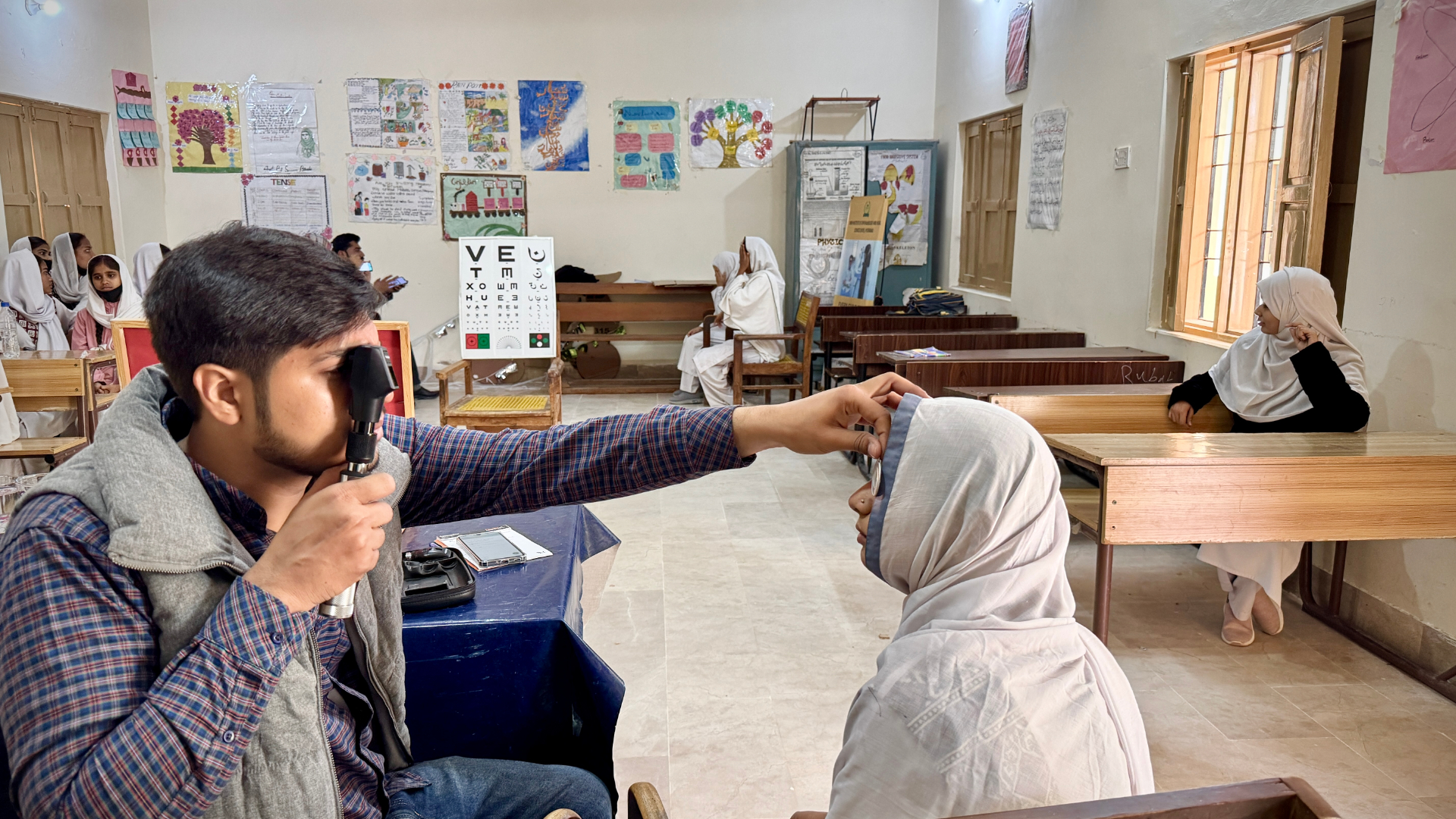 Optometrist performs retinoscopy on a student in a Pakistani classroom with vision chart on the wall
