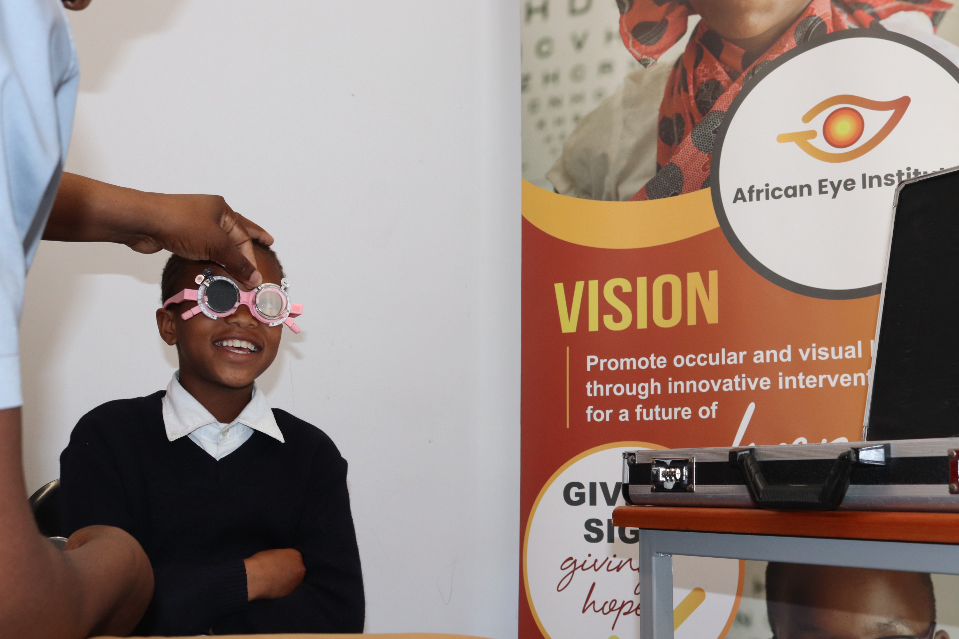A learner from Umyezo Wama Apile Primary smiling while wearing a pink trial frame, with the African Eye Institute banner in the background