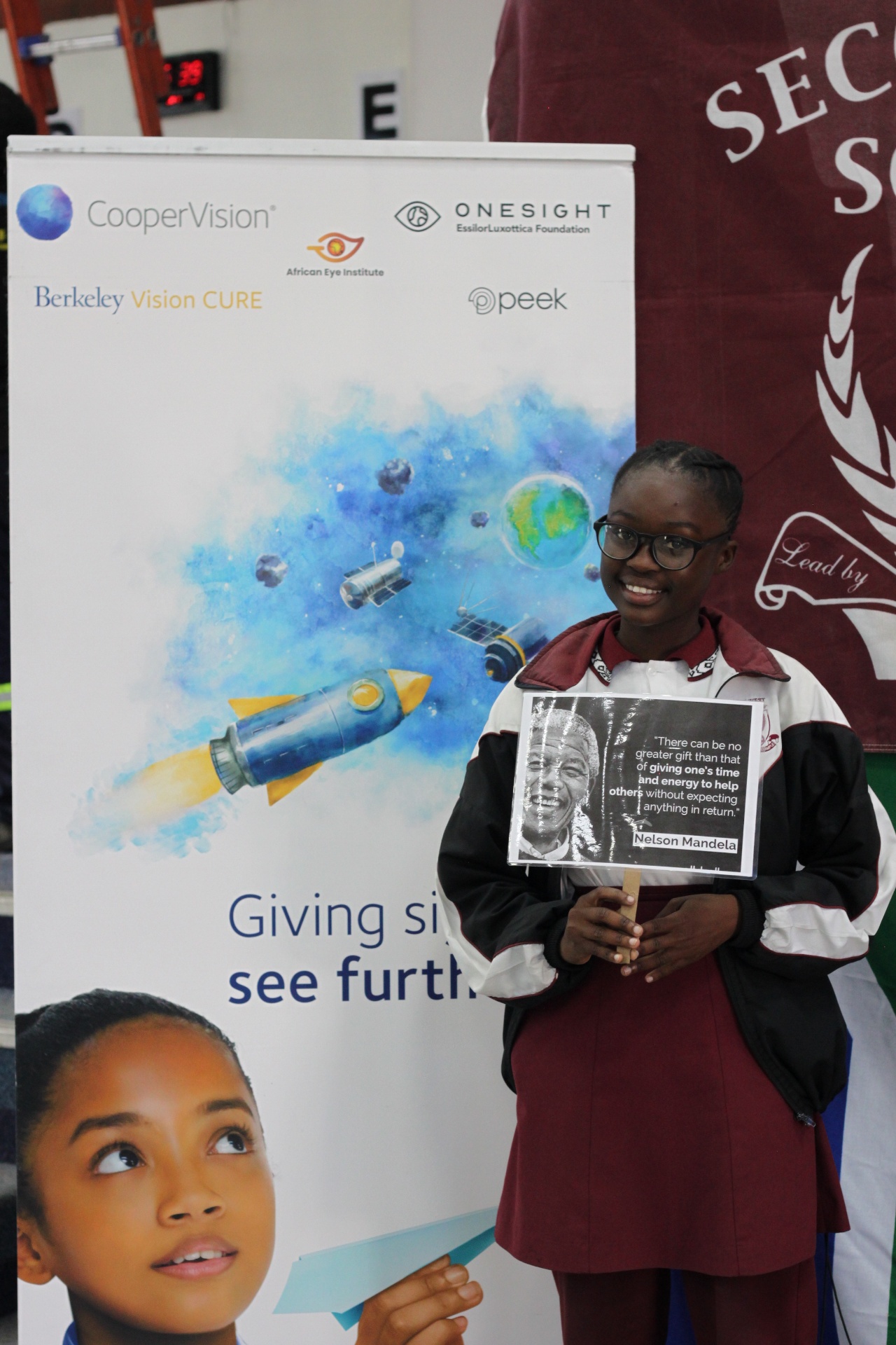 Student in new glasses holds a Nelson Mandela quote at a partner-branded backdrop