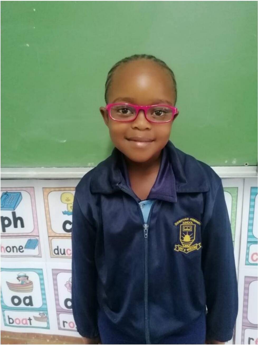 Syamthanda, a young girl wearing pink glasses and her Sunnyvale Primary uniform, smiling in front of a classroom chalkboard